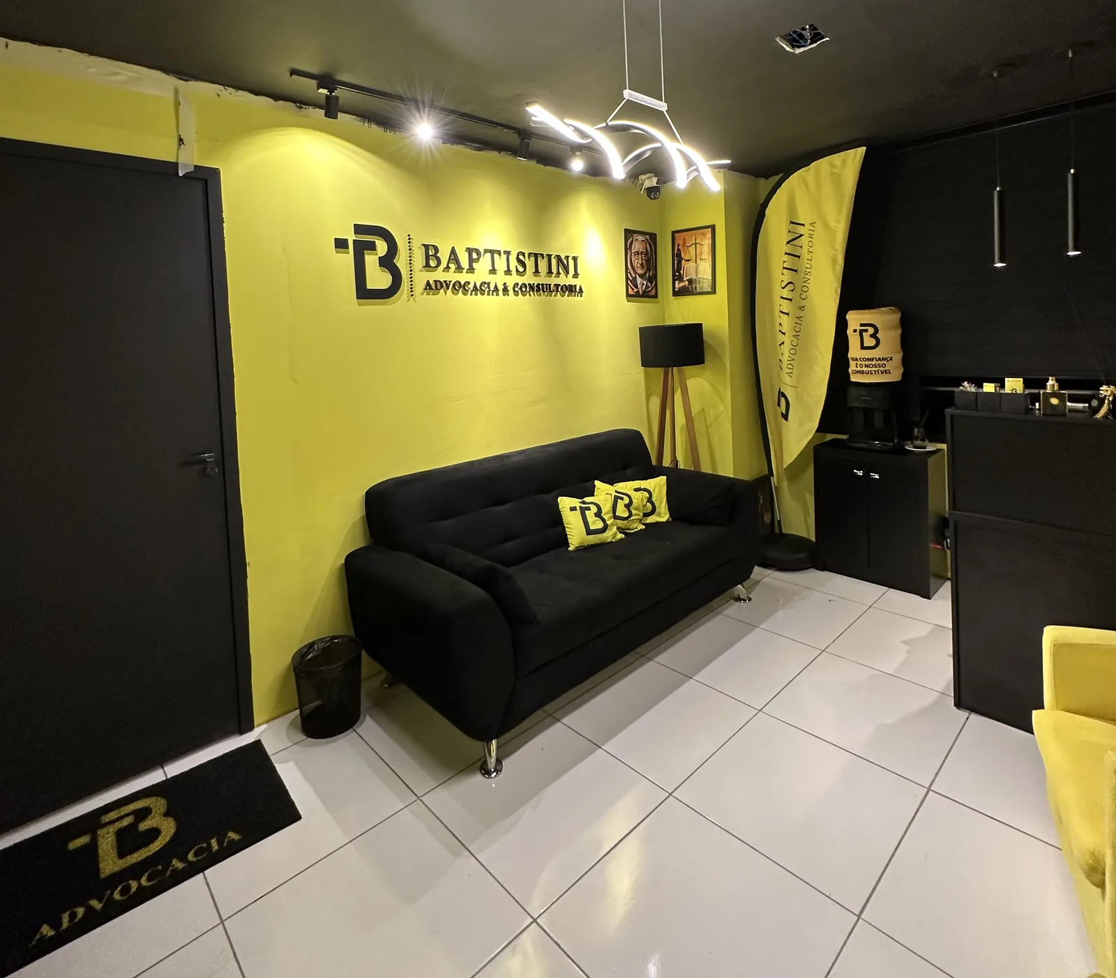 Recepção do escritório: sofá preto, parede amarela e a marca Baptistini iluminada